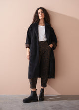 Woman wearing a long black fur coat, white top, and leopard print trousers against a beige wall.