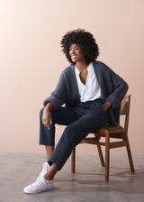 Woman sitting on a chair wearing a cardigan, white shirt, and dark grey jersey trousers against a beige background