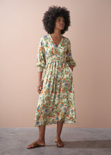 A woman in a long, floral, v-neck satin dress, and brown leather sandals against a beige wall.