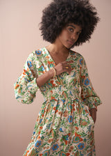 Close-up of a woman in a long, floral, v-neck satin dress, with her hand tucked into the pocket, against a beige wall.