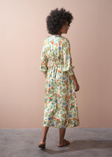 A woman in a long, floral, v-neck satin dress, and brown leather sandals, facing toward a beige wall.