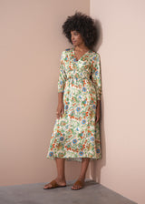 A woman in a long, floral, v-neck satin dress, and brown leather sandals, leaning against a beige wall.