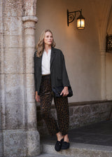 A woman stands on stone steps in a historic building, wearing a dark charcoal blazer, white blouse, leopard-print trousers, and black loafers, holding a black clutch.