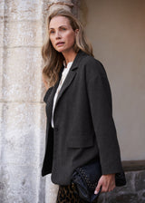 A woman wearing an oversized dark charcoal gray blazer over a white top, standing outdoors next to a stone column and holding a black studded clutch.