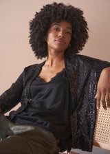 Woman wearing a black sequin jacket over a black top, sitting on a beige chair.