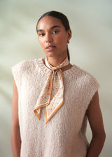 Woman wearing a beige knitted sleeveless top with a floral scarf around her neck against a plain background
