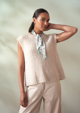 Woman wearing a beige knitted vest and light-colored pants with a green scarf around her neck against a plain background