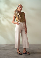 Woman wearing a mustard top, beige scarf, and white pants against a neutral background