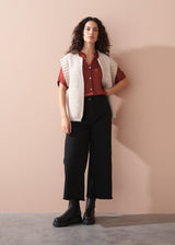 Woman wearing a white sleeveless cardigan over a red shirt with black trousers against a beige wall.