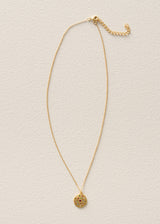 A delicate gold chain necklace with a small circular textured pendant, displayed on a textured off-white background.