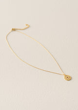 A delicate gold chain necklace with a small, textured circular pendant, displayed on a light beige textured background.