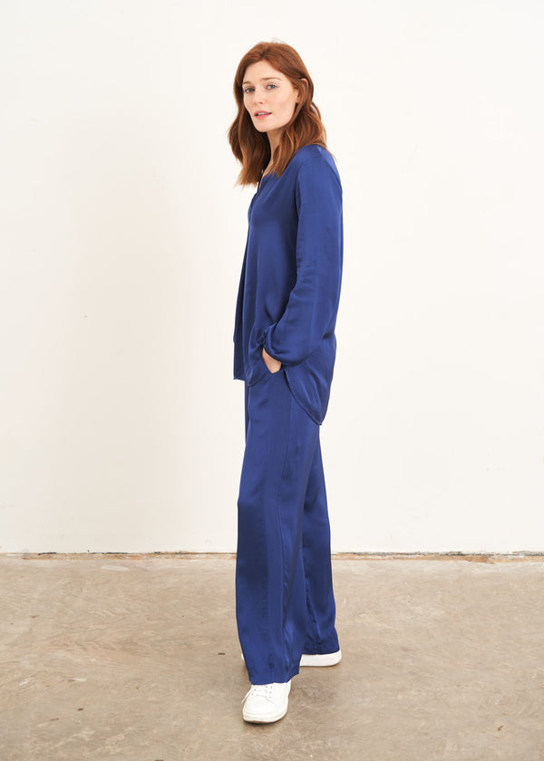 A model wearing a navy blue satin co-ord consisting of a long sleeved blouse with a v neck and wide leg long trousers