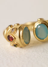 A gold open ring with two decorative ends featuring light aqua semi precious oval stones and small dark brown gem accents, photographed on a textured off-white background