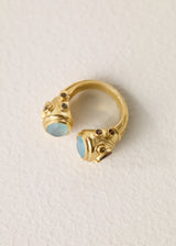 A gold open ring with two decorative ends featuring light aqua semi precious oval stones and small dark brown gem accents, photographed on a textured off-white background