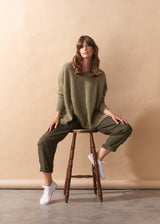 Woman sitting on a wooden stool wearing a khaki green wool blend sweater and pants against a beige background