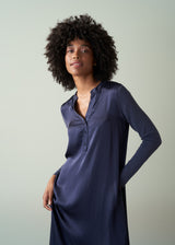 Woman in a long, dark navy satin v-neck dress, standing against a plain green background