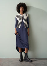 A woman in a long, dark navy satin dress, striped knitted waistcoat, and black leather boots leaning against a green wall.