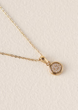 A delicate gold chain necklace with a small round bezel-set pendant paved with clear stones, displayed on a textured off-white background.