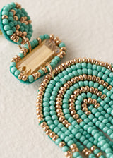 A pair of beaded tassel earrings featuring turquoise and gold seed beads in a semi-circular arch pattern with dangling fringe, displayed on a textured off-white background.