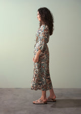 Woman in an abstract print satin midi dress with detachable belt, facing left, and standing against a green wall