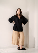 Woman wearing a black top and beige pants against a white wall