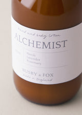 Close up of amber bottle of hand lotion, featuring "Alchemist" branding, on a beige background