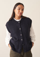 Woman wearing a dark navy blue sleeveless chunky knit waistcoat with green trousers and a white shirt