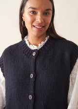 Woman wearing a dark navy blue sleeveless chunky knit waistcoat with green trousers and a white shirt
