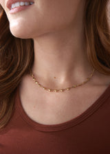 A woman wearing a gold necklace with heart-shaped charms, styled with a brown jersey vest.