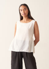 A woman wearing a sleeveless white top with a relaxed, slightly sheer texture and a peplum-like hem, paired with dark wide-leg trousers.