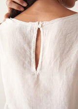 A woman wearing a sleeveless white top with a relaxed, slightly sheer texture and a peplum-like hem, paired with dark wide-leg trousers.