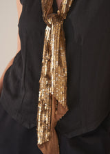 Woman wearing black waistcoat and gold sequin scarf against a plain background.