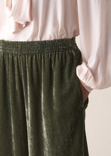 Woman wearing green velvet cropped wide-leg trousers and a pink shirt against a plain background