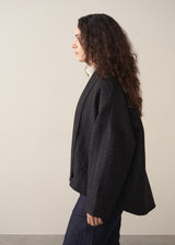 Woman wearing a black quilted kimono jacket over a beige sweater and dark jeans against a plain background