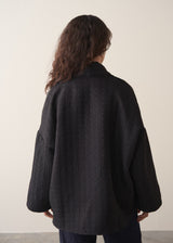 Woman wearing a black quilted kimono jacket over a beige sweater and dark jeans against a plain background