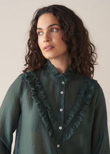 Woman wearing dark green ruffle front shirt, set against a beige background