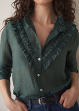 Woman wearing dark green ruffle front shirt with belted blue jeans against a beige background
