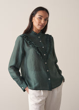 Woman wearing a dark green blouse with ruffled details and cream suit trousers.