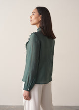 Woman wearing a dark green blouse with ruffled details and cream suit trousers.
