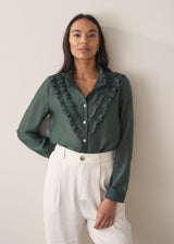 Woman wearing a dark green blouse with ruffled details and cream suit trousers.