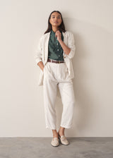 Woman wearing a dark green blouse with ruffled details, cream suit trousers and white loafers against a beige background.
