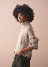 Person wearing a beige chunk knit fisherman sweater and green barrel leg trousers against a beige background