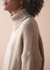 Woman wearing a beige chunky knit fisherman sweater and green barrel leg trousers with a beige background