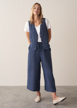 Woman wearing a blue linen waistcoat over a v-neck white tee with wide leg linen trousers and loafers against a plain background