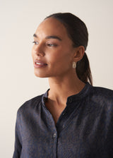 Model wearing gold drop stud earrings with delicate white beads, paired with a blue leopard print shirt, against a neutral beige background