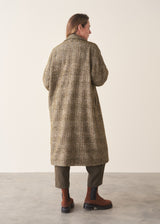 A woman wearing an oversized checked coat with dark green trousers, ginger boots and a white top against a plain background.