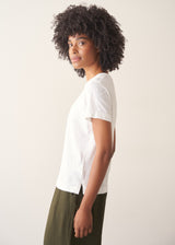 Woman wearing white, round neck t-shirt and dark green wide leg trousers against a plain background