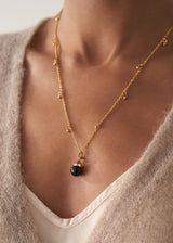 Model wears a long golden chain necklace with gold beads and a black pendant