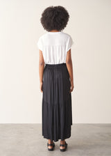 Woman wearing a white top and black satin tiered midi skirt against a plain background