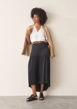 Woman wearing a white top and black satin tiered midi skirt with beige blazer against a plain background